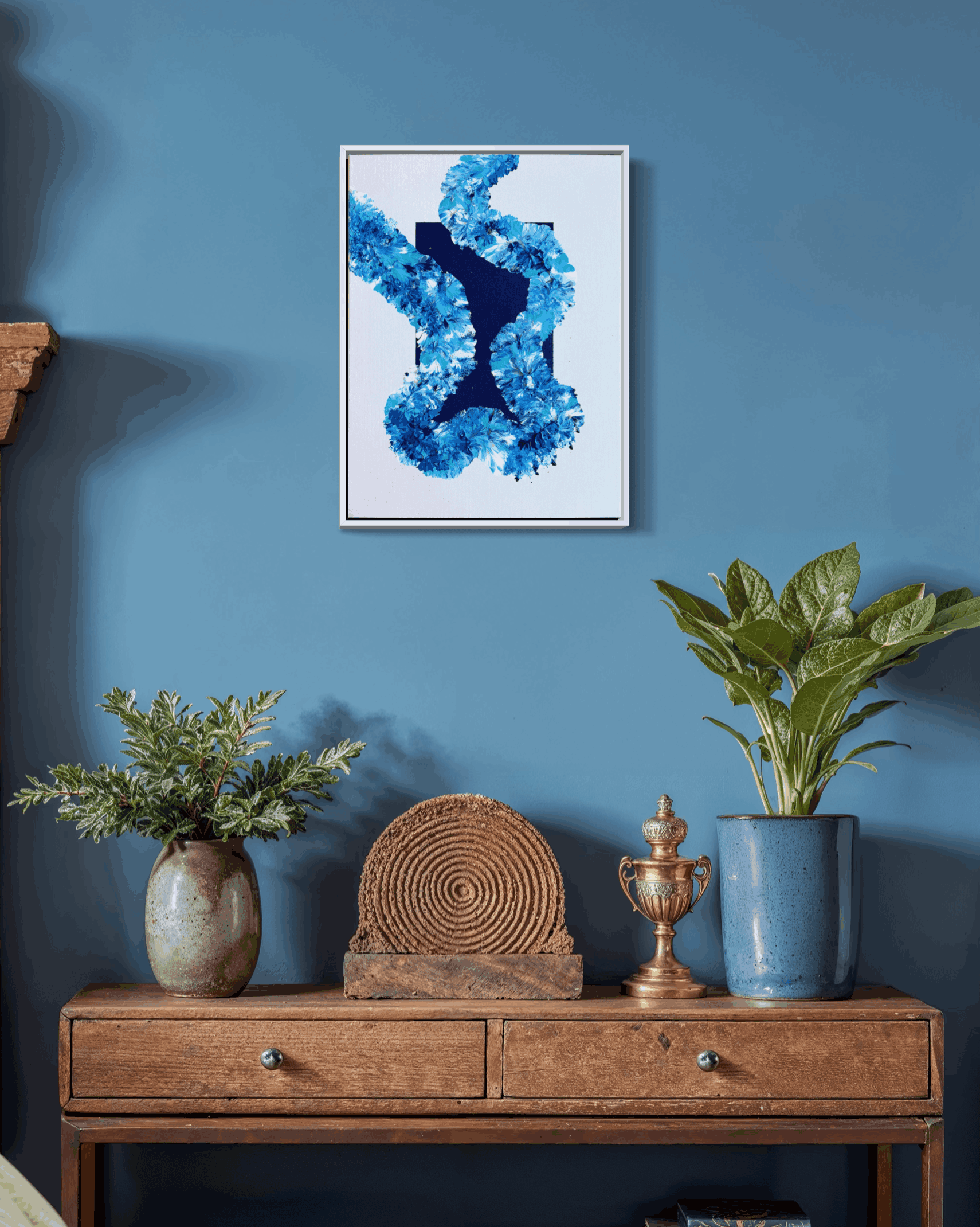 A framed abstract art print featuring a blue and white design, displayed on a blue wall above a rustic wooden console table with plants and decorative objects.