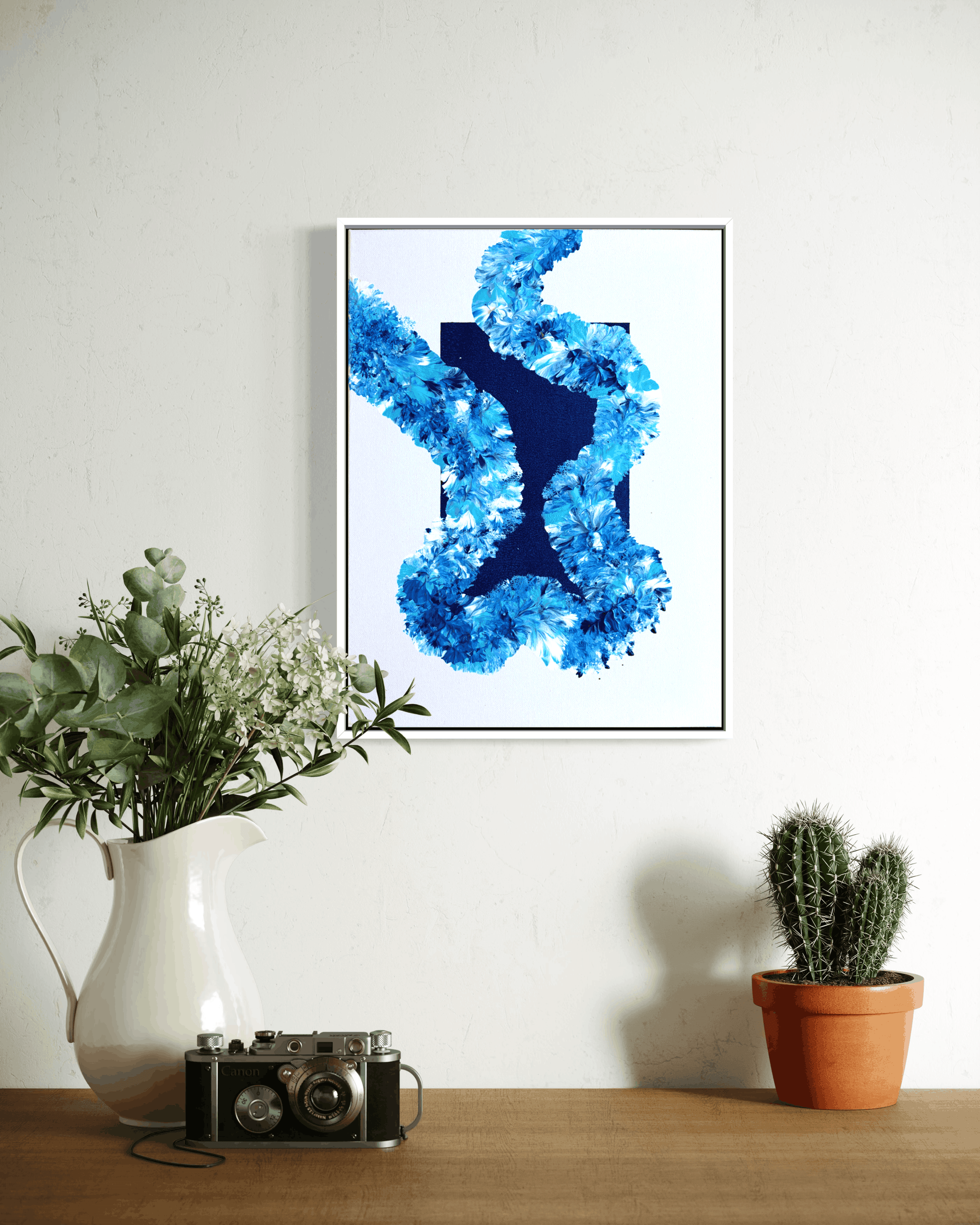 A framed abstract art print featuring a textured blue and navy blue design on a white background, hanging on a white wall above a wooden table styled with a white pitcher of greenery, a vintage camera, and a small potted cactus.