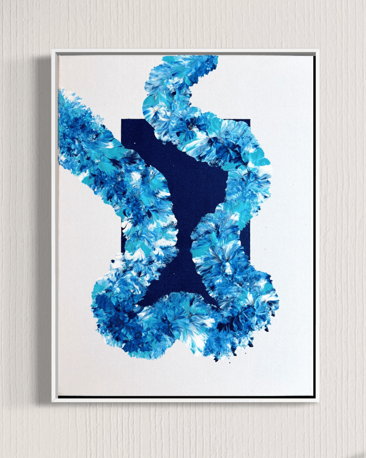 A framed abstract art print featuring a textured blue floral or coral-like pattern around a dark navy X-shaped center, mounted on a light beige textured wall.