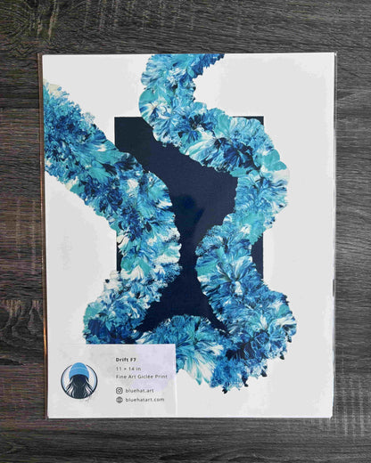 An art print titled 'Draft 2' featuring a white background with a large blue and teal floral or feather-like graphic shape, photographed on a dark wood surface. A small label at the bottom left shows the artist name 'Skeletart' and website 'skelartart.