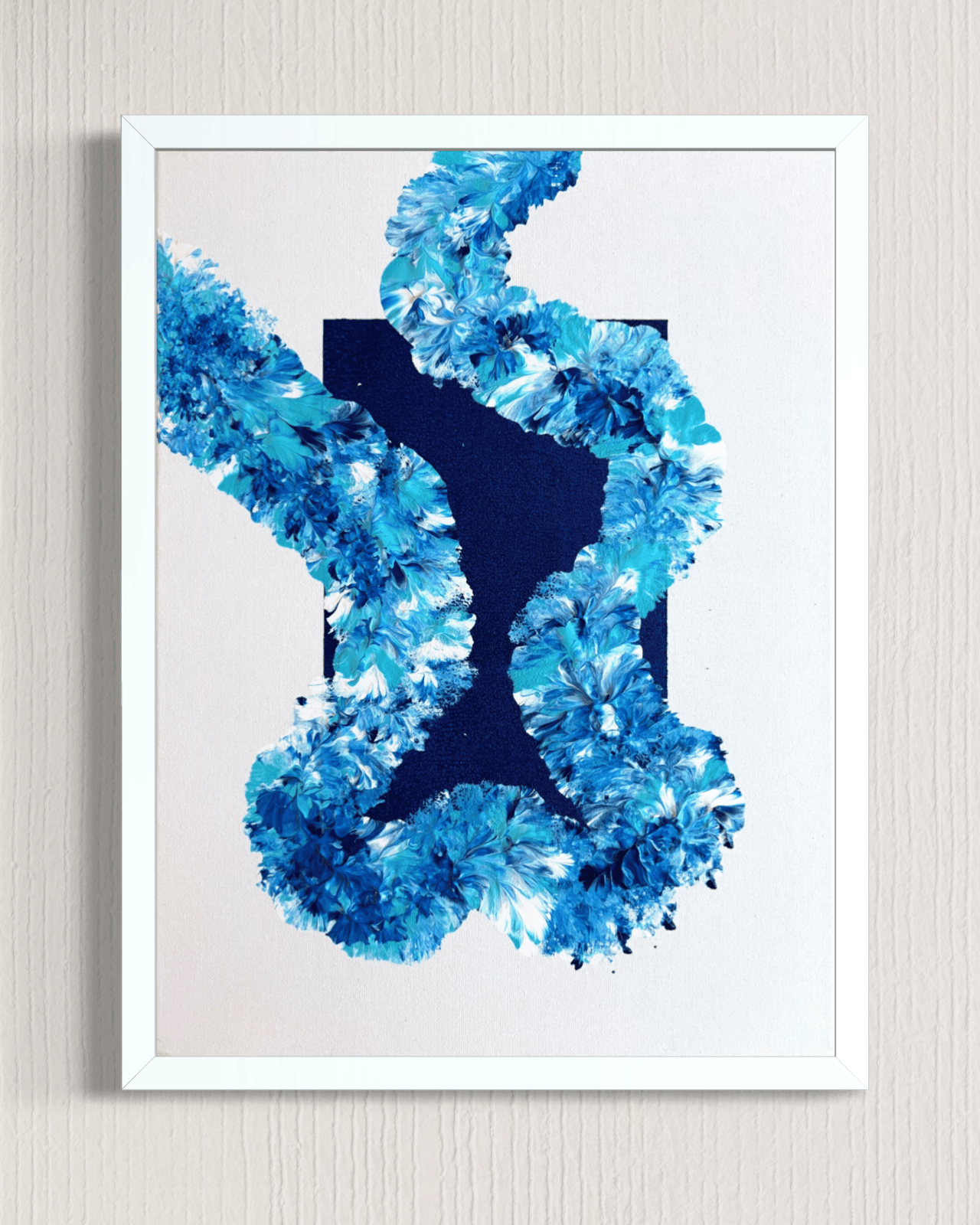 A framed abstract art print featuring a textured blue floral or coral-like pattern around a dark navy central shape, set against a white background and mounted on a light beige wall.
