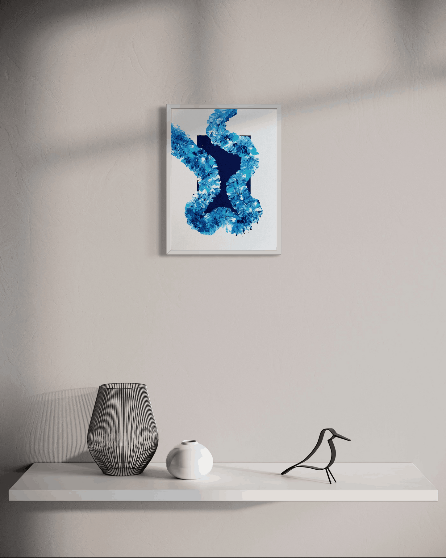 A framed abstract art print featuring textured blue and navy shapes on a white background, displayed on a light gray wall above a white shelf with decorative objects.