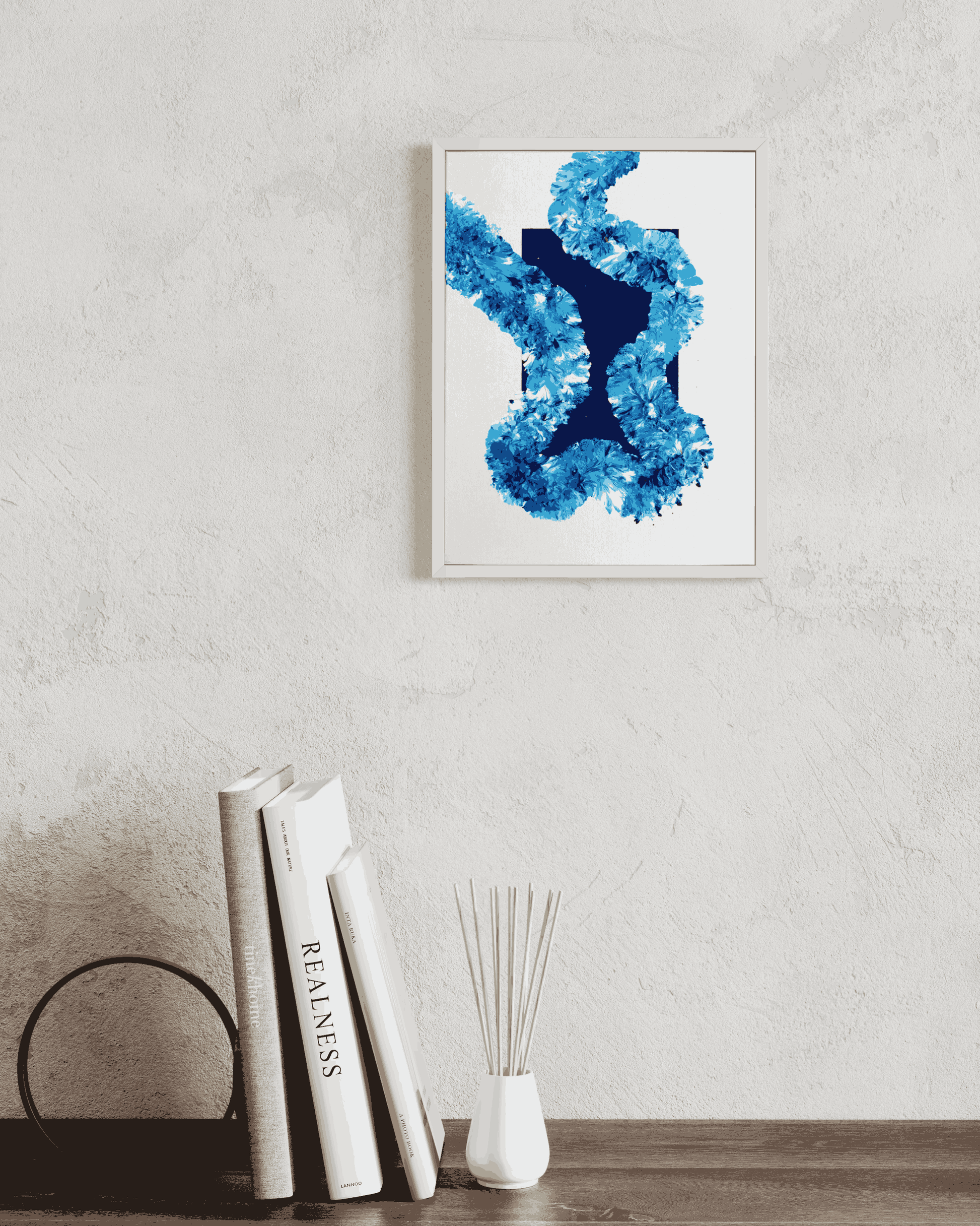 A framed abstract art print featuring a textured blue and navy brushstroke design on a white background, hanging on a textured light gray wall above a wooden shelf with books and a reed diffuser.