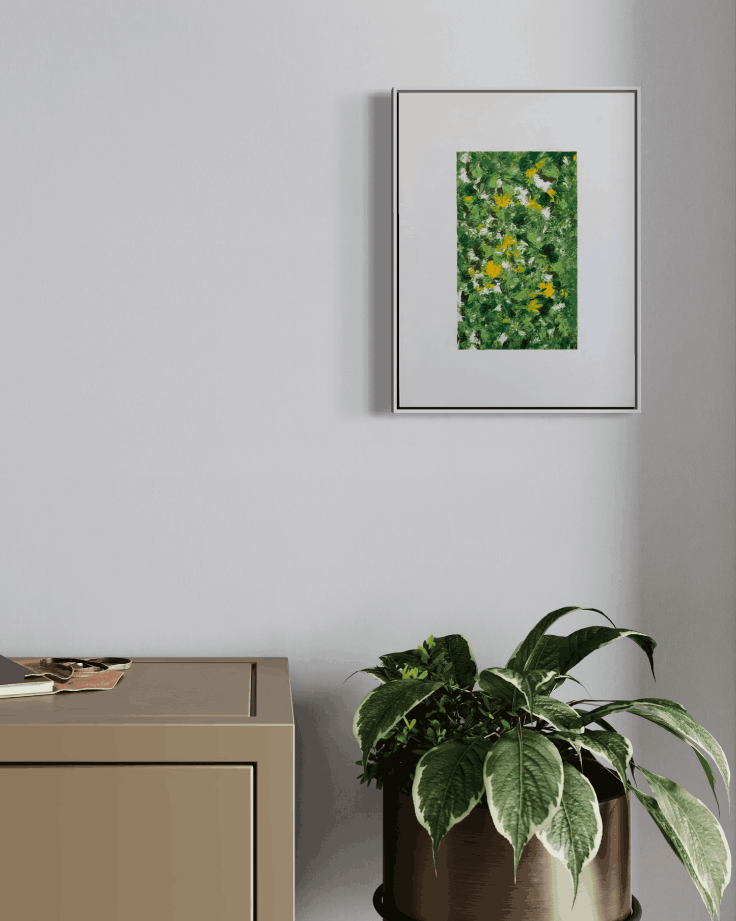 A framed abstract art print featuring green foliage with small yellow and white flowers, displayed on a light gray wall above a beige cabinet and next to a potted green plant.