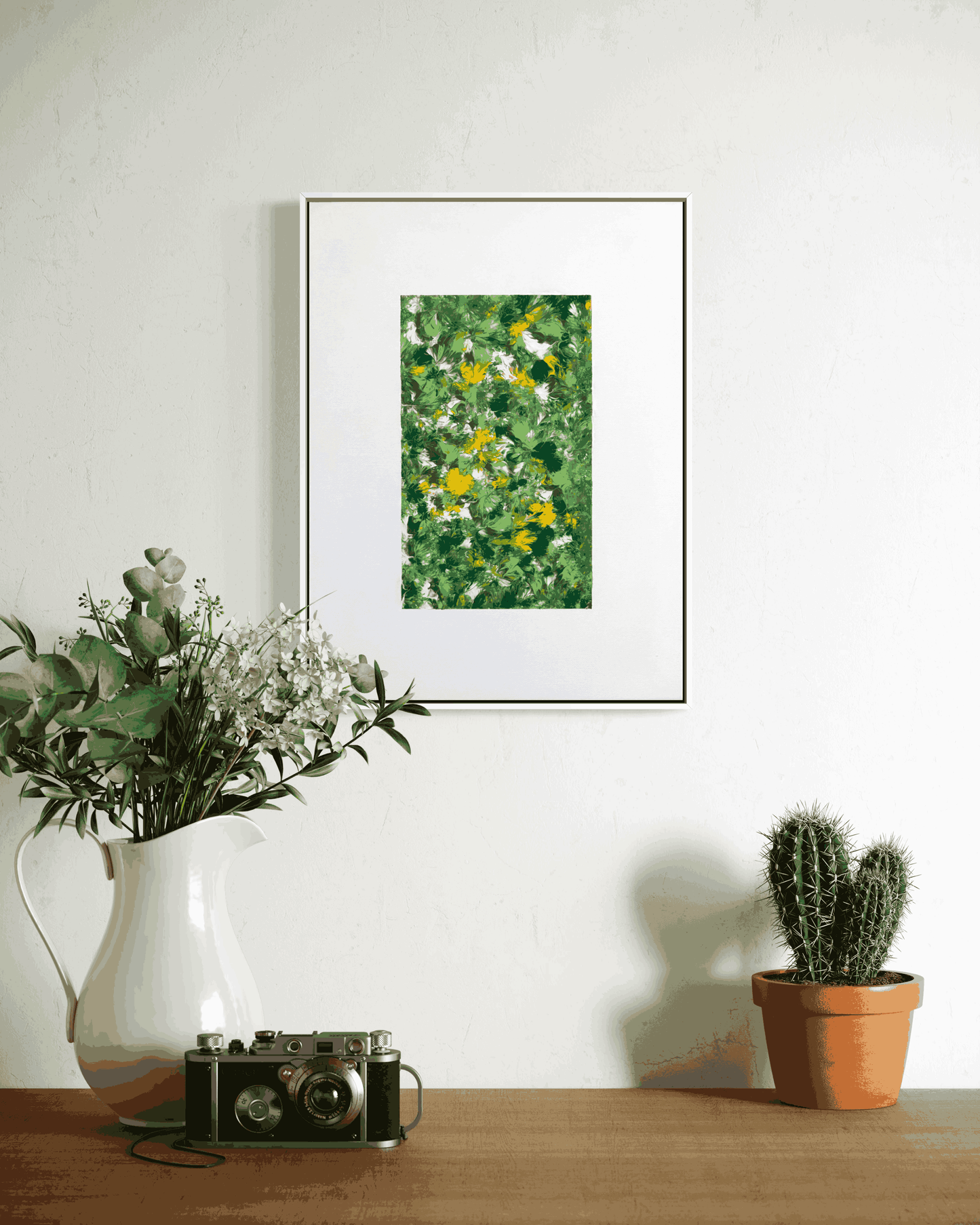 A framed art print featuring a green and yellow textured floral pattern, displayed on a white wall above a wooden table styled with a white vase of flowers, a vintage camera, and a small potted cactus.