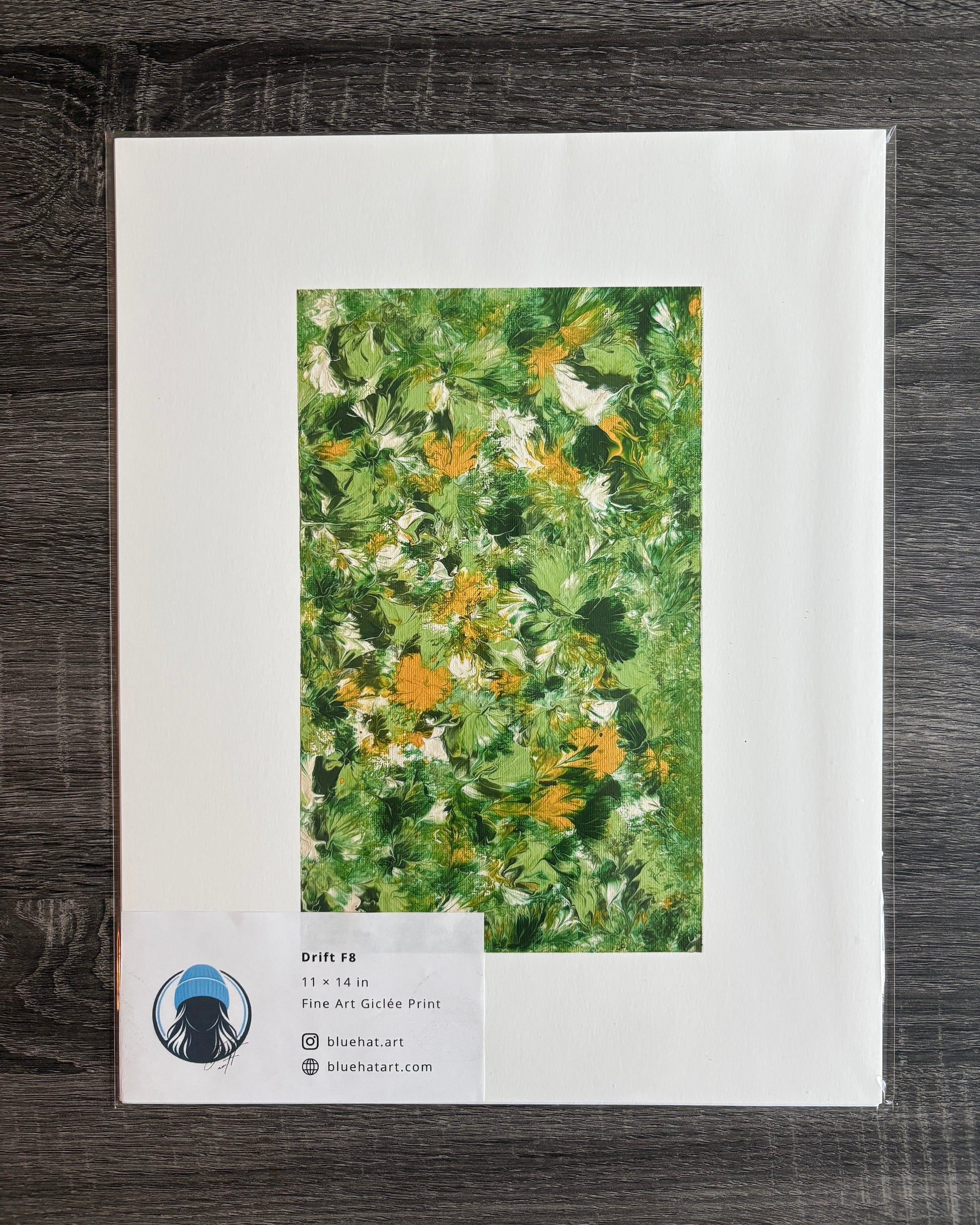 A flat-lay photograph of a matted art print on a dark wood background. The print features a textured green and yellow floral painting surrounded by a wide white mat border.