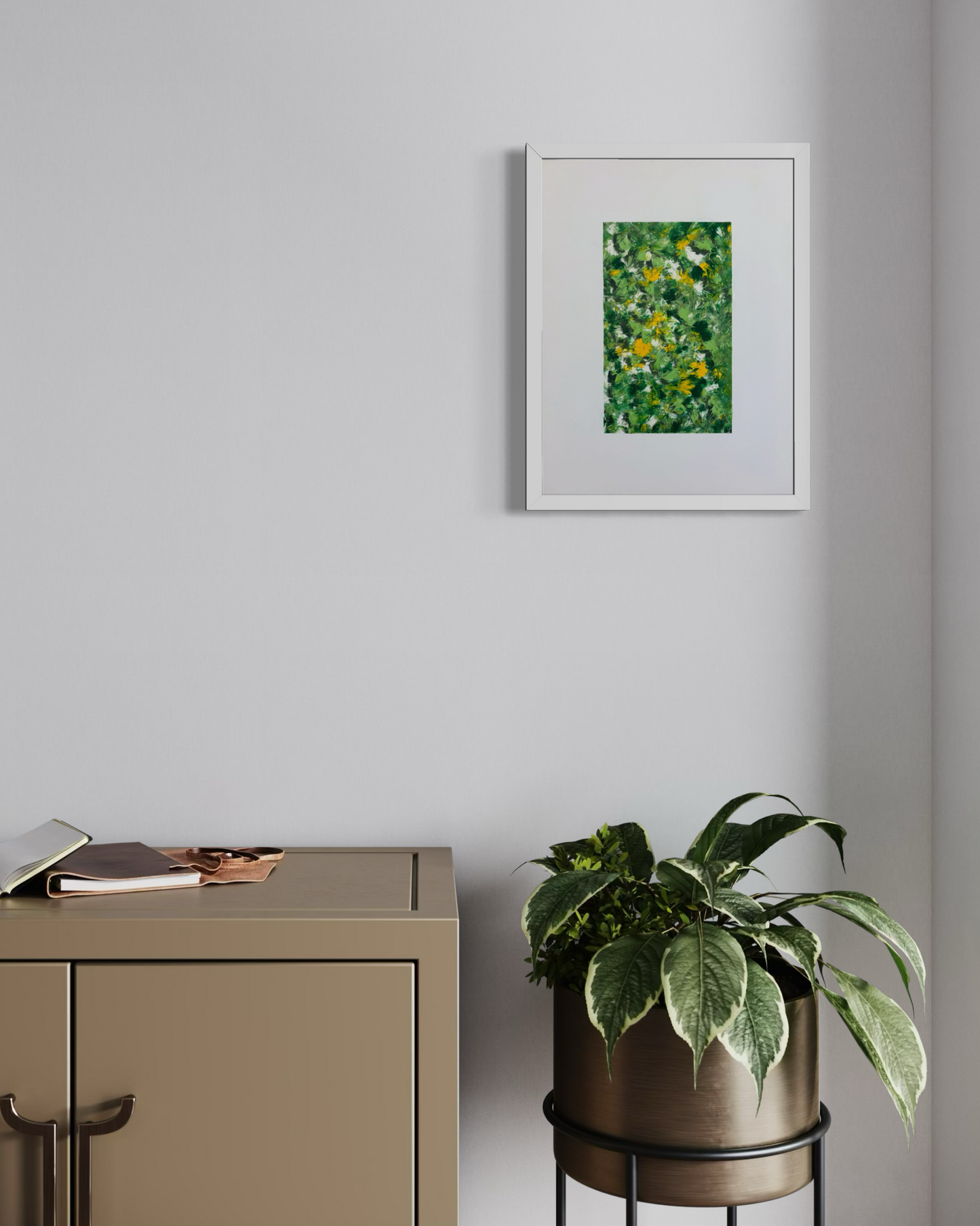 A framed abstract green and yellow painting hangs on a light gray wall above a beige cabinet, with a potted plant on a black metal stand nearby.