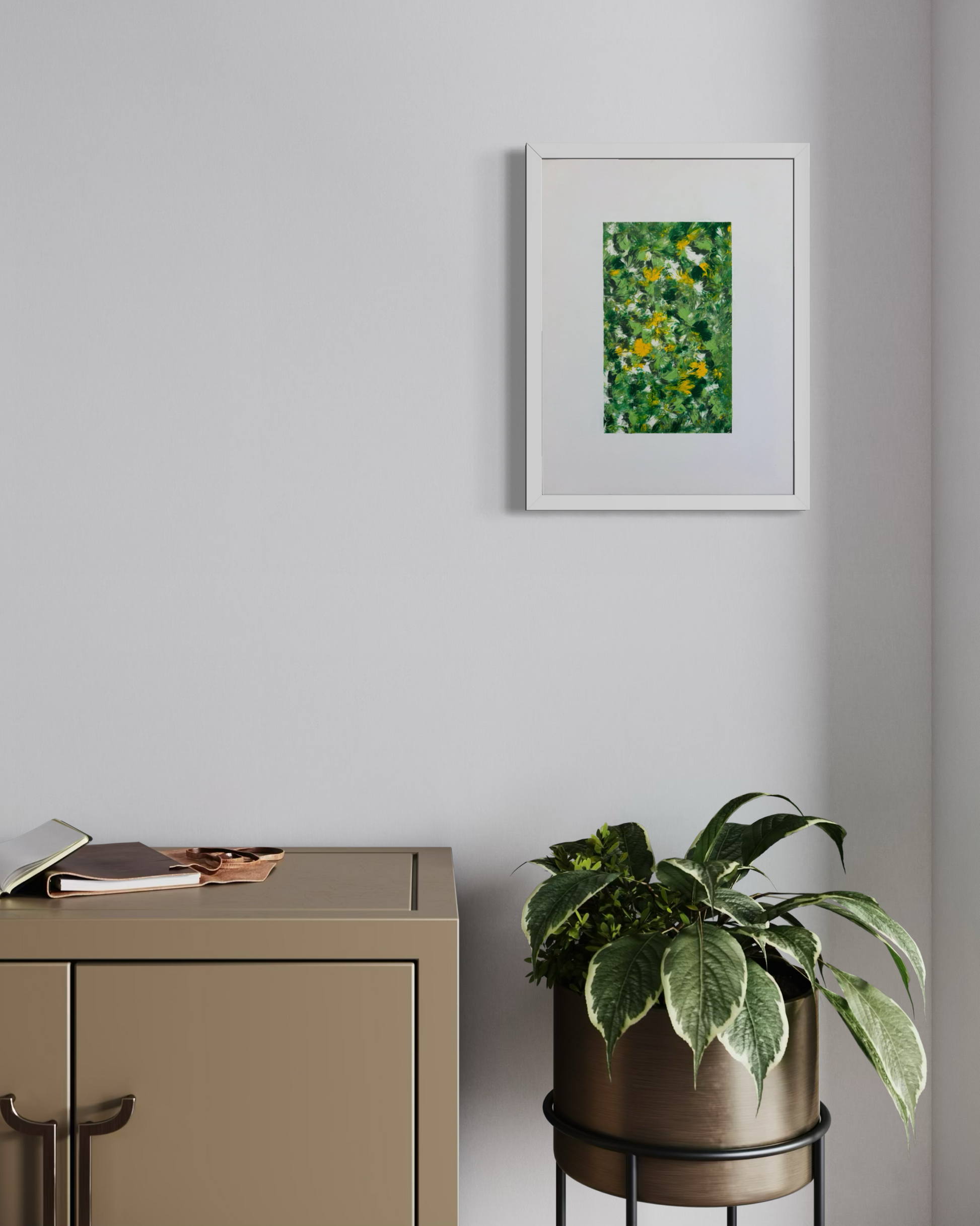 A framed abstract green and yellow painting hangs on a light gray wall above a beige cabinet, with a potted plant on a black metal stand nearby.
