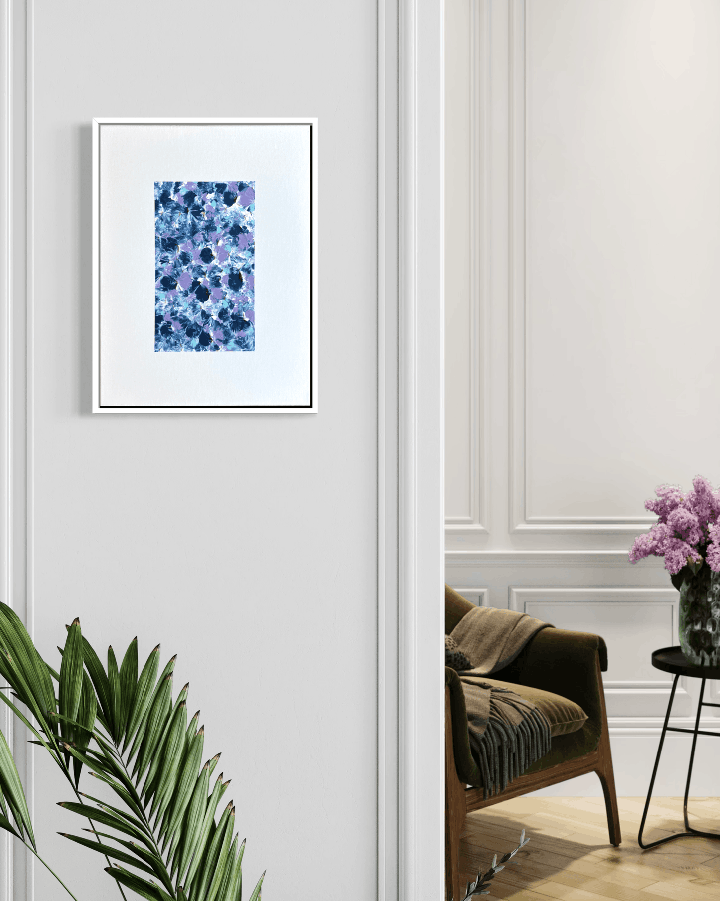A framed abstract floral art print in shades of blue and purple, displayed on a light gray paneled wall in a styled interior room with a chair, side table, and plant.