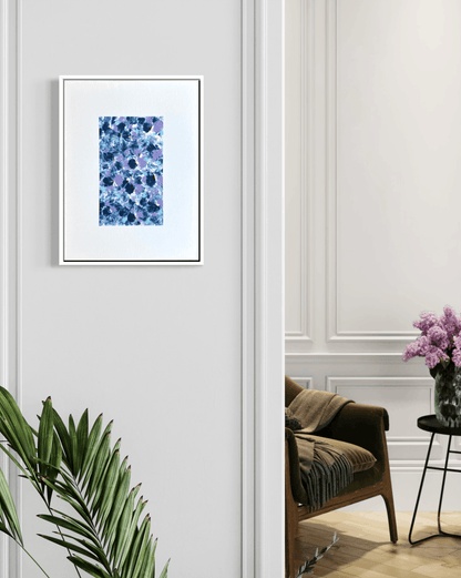 A framed abstract floral art print in shades of blue and purple, displayed on a light gray paneled wall in a styled interior room with a chair, side table, and plant.