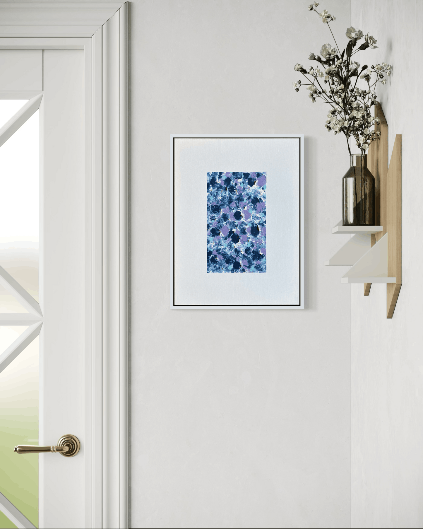 A framed abstract art print featuring blue and purple watercolor-like splashes, displayed on a white wall next to a wooden shelf with dried flowers.