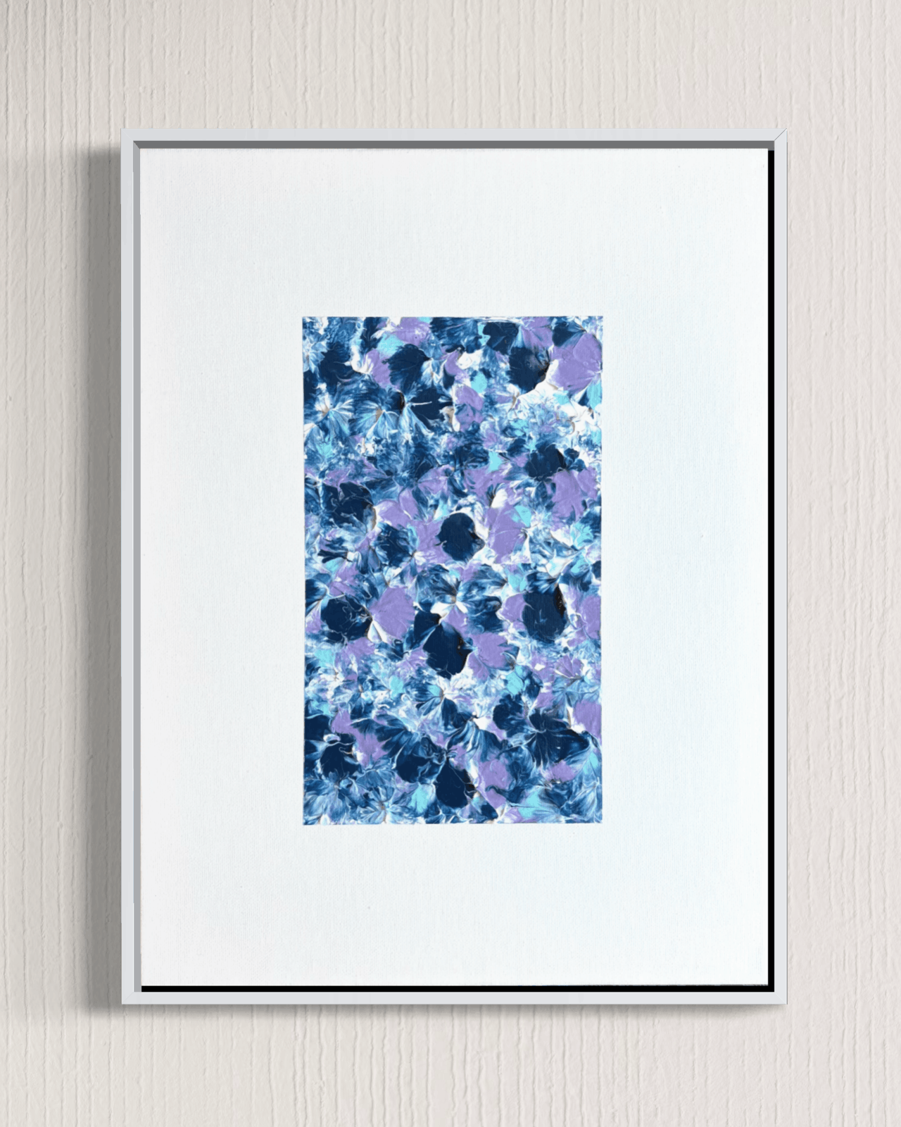 A framed abstract floral art print featuring blue, purple, and navy watercolor-like blossoms, presented in a white mat and thin black frame against a light textured wall.
