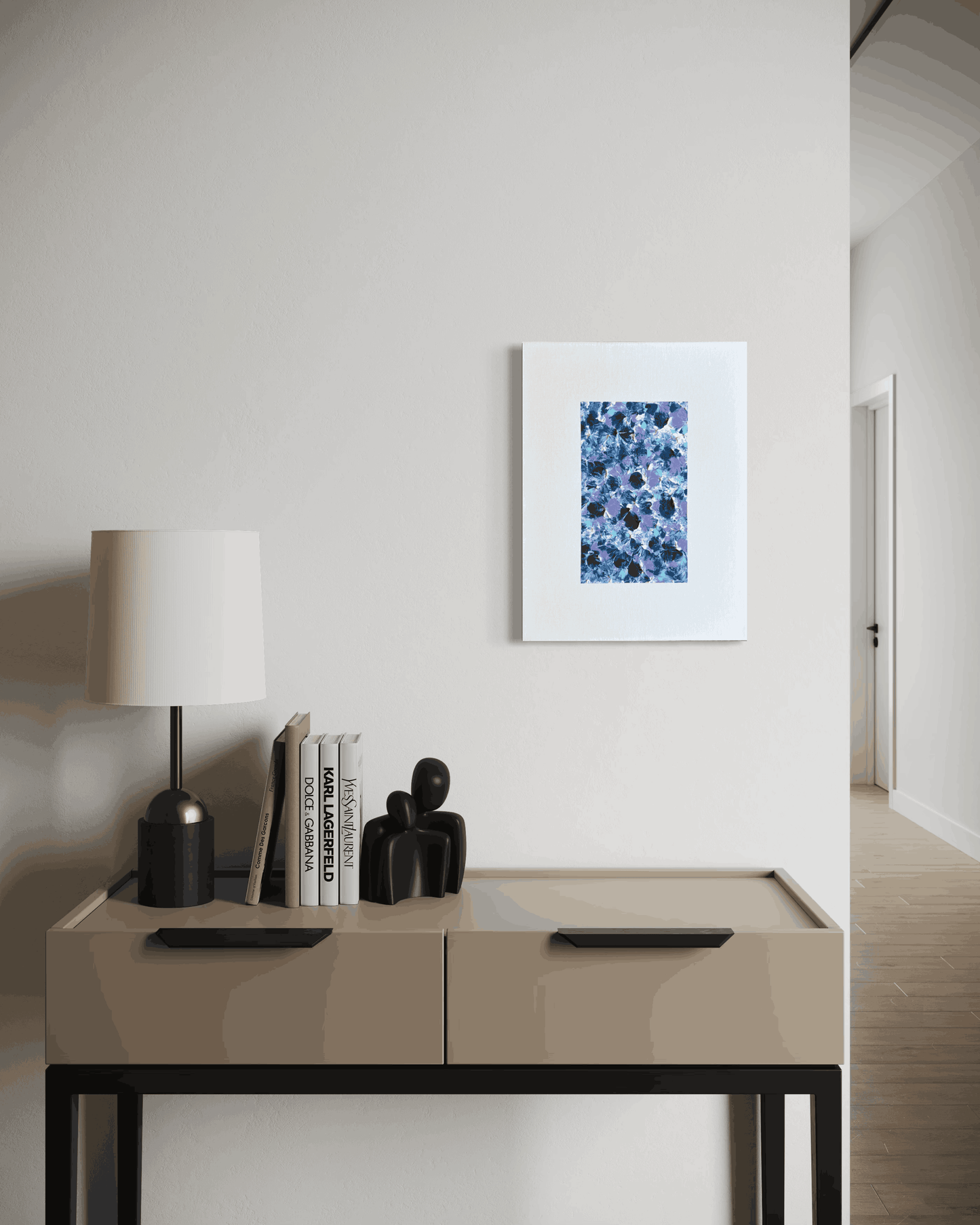 A framed abstract art print featuring a blue and white floral pattern, displayed on a light beige wall above a modern console table with a lamp and books.