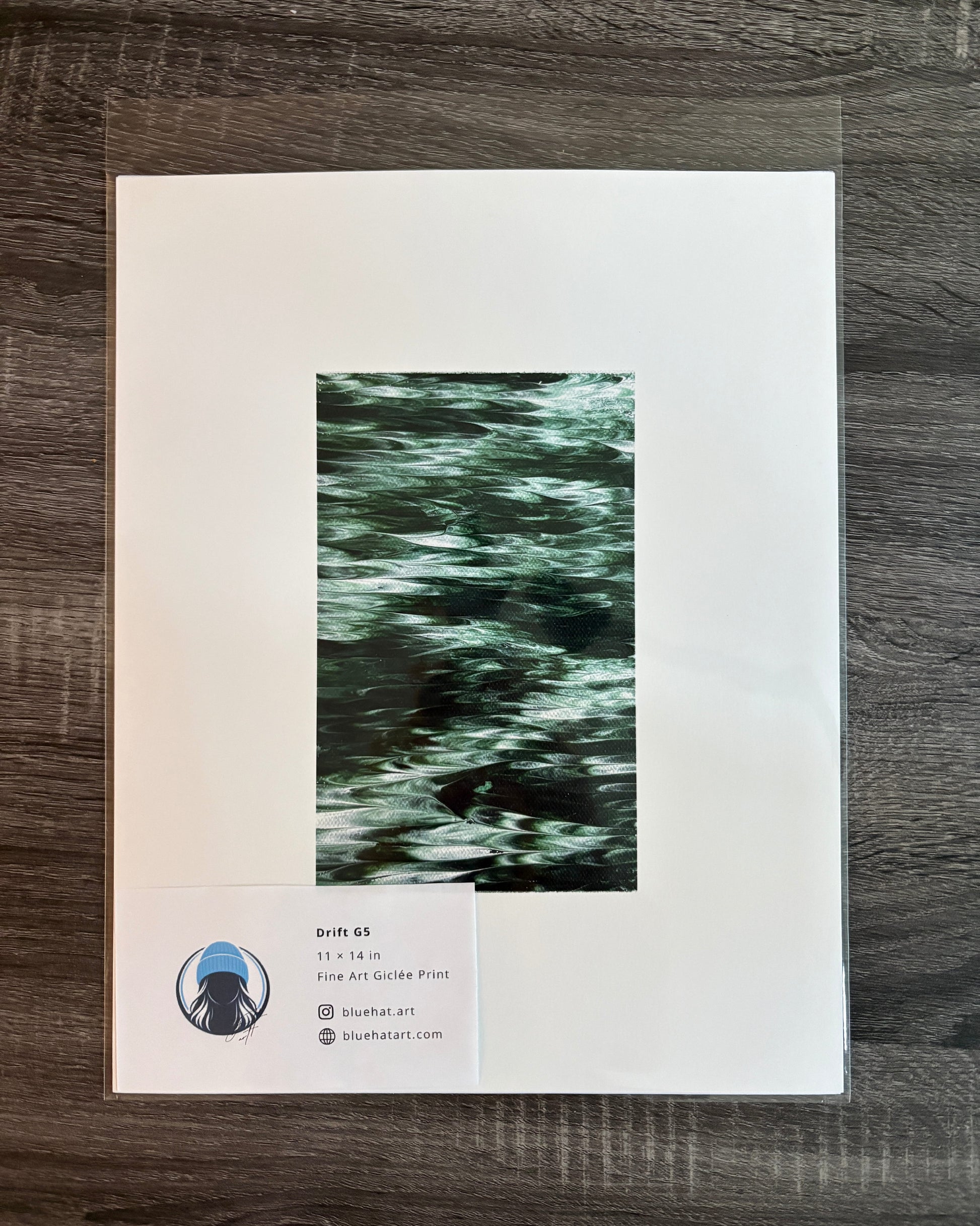 A framed or matted art print laid flat on a dark wood surface. The print has a wide white border and a central vertical artwork in dark green and black tones.