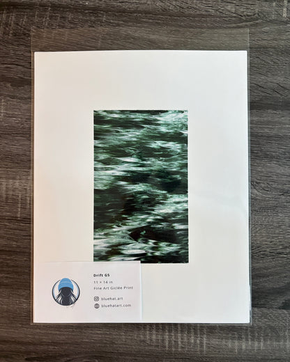 A framed or matted art print laid flat on a dark wood surface. The print has a wide white border and a central vertical artwork in dark green and black tones.