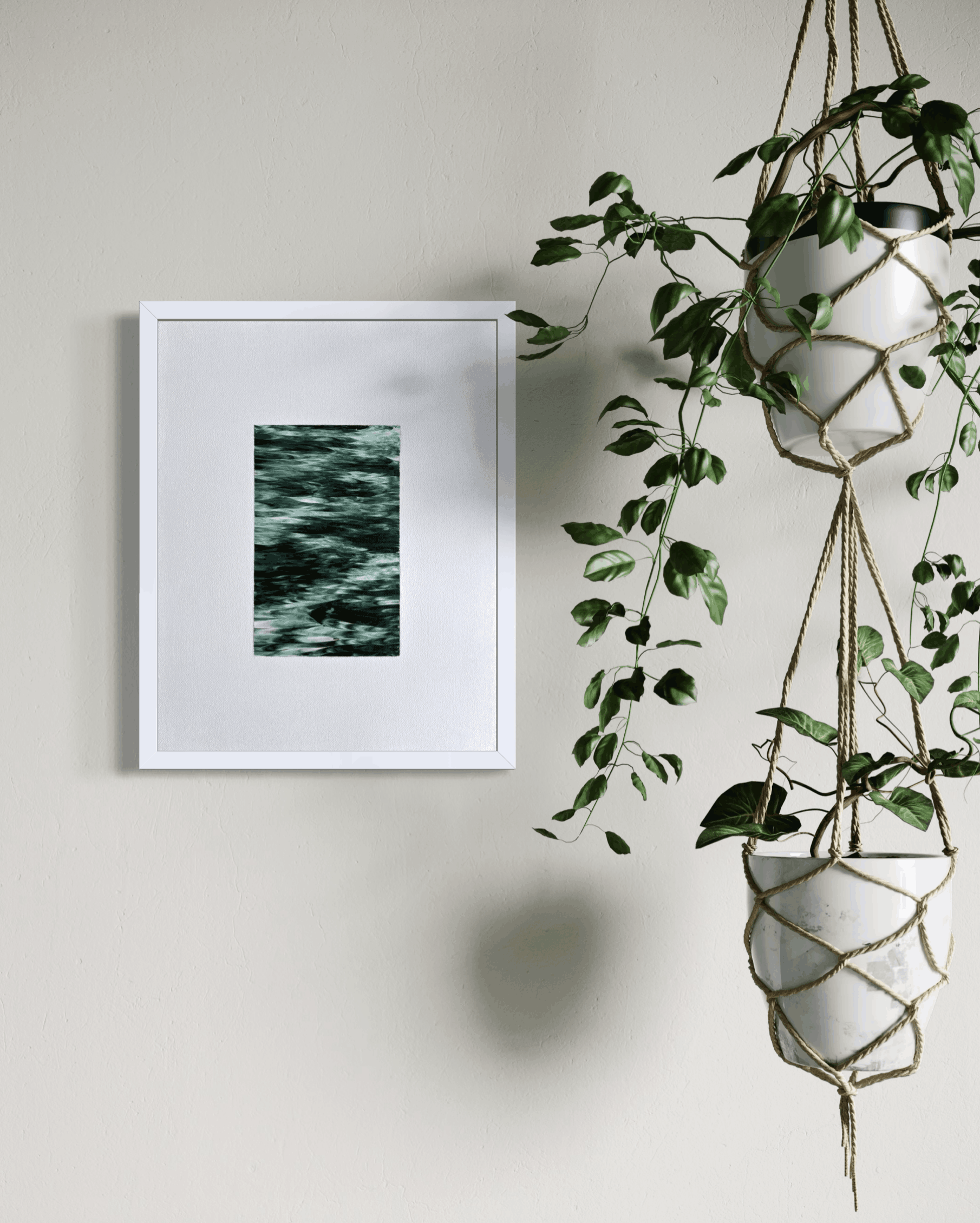 A white square frame containing a small abstract green and dark teal print, hung on a light beige wall next to a hanging green plant in a white pot with a rope hanger.