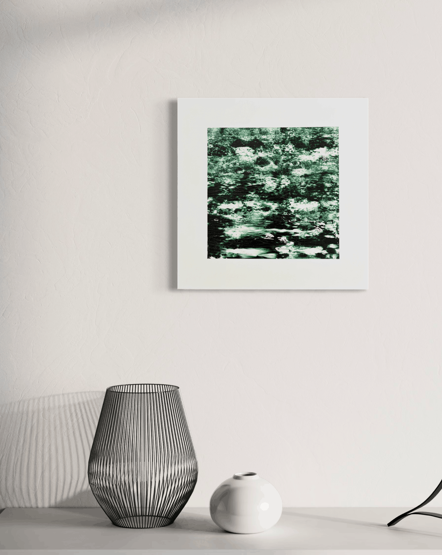 A square framed art print featuring a green and white textured pattern, displayed on a white wall above a shelf with decorative objects.