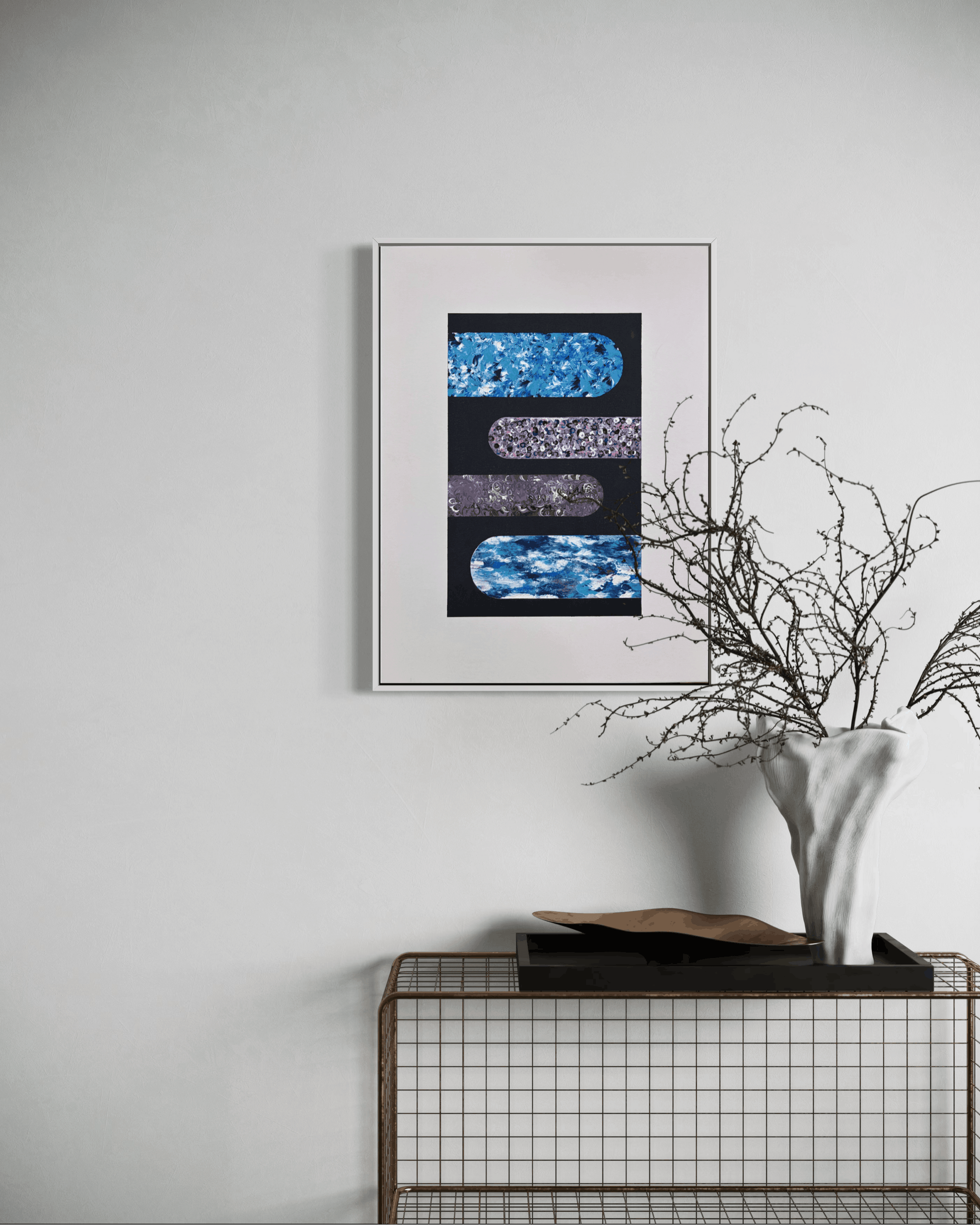 A framed abstract art print featuring textured blue and purple horizontal shapes on a black background, styled on a light gray wall above a wire grid console table with a white vase of branches.