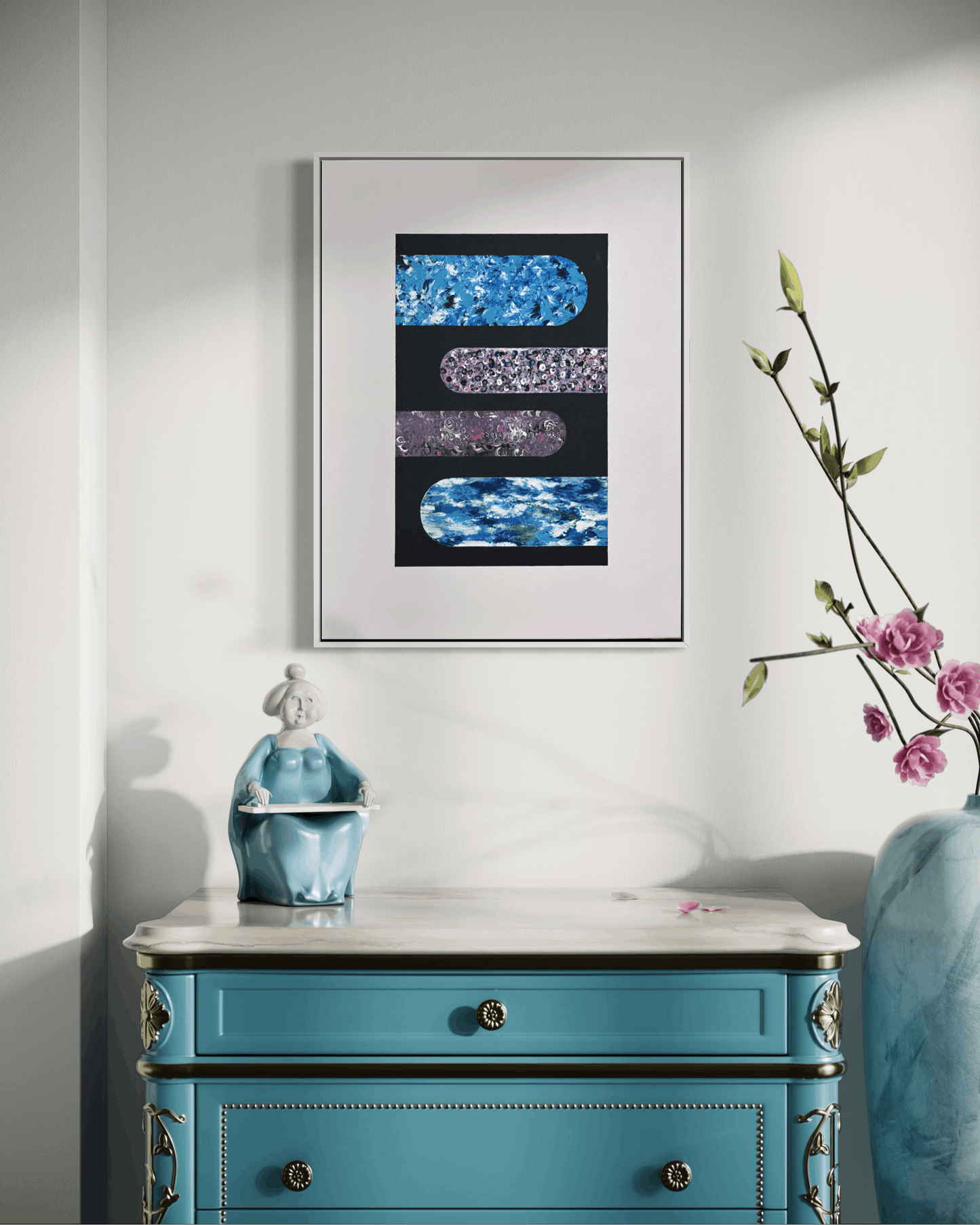 A framed abstract art print featuring textured blue, purple, and silver horizontal bars on a black background, styled in a room above a turquoise console table with a vase of pink flowers.