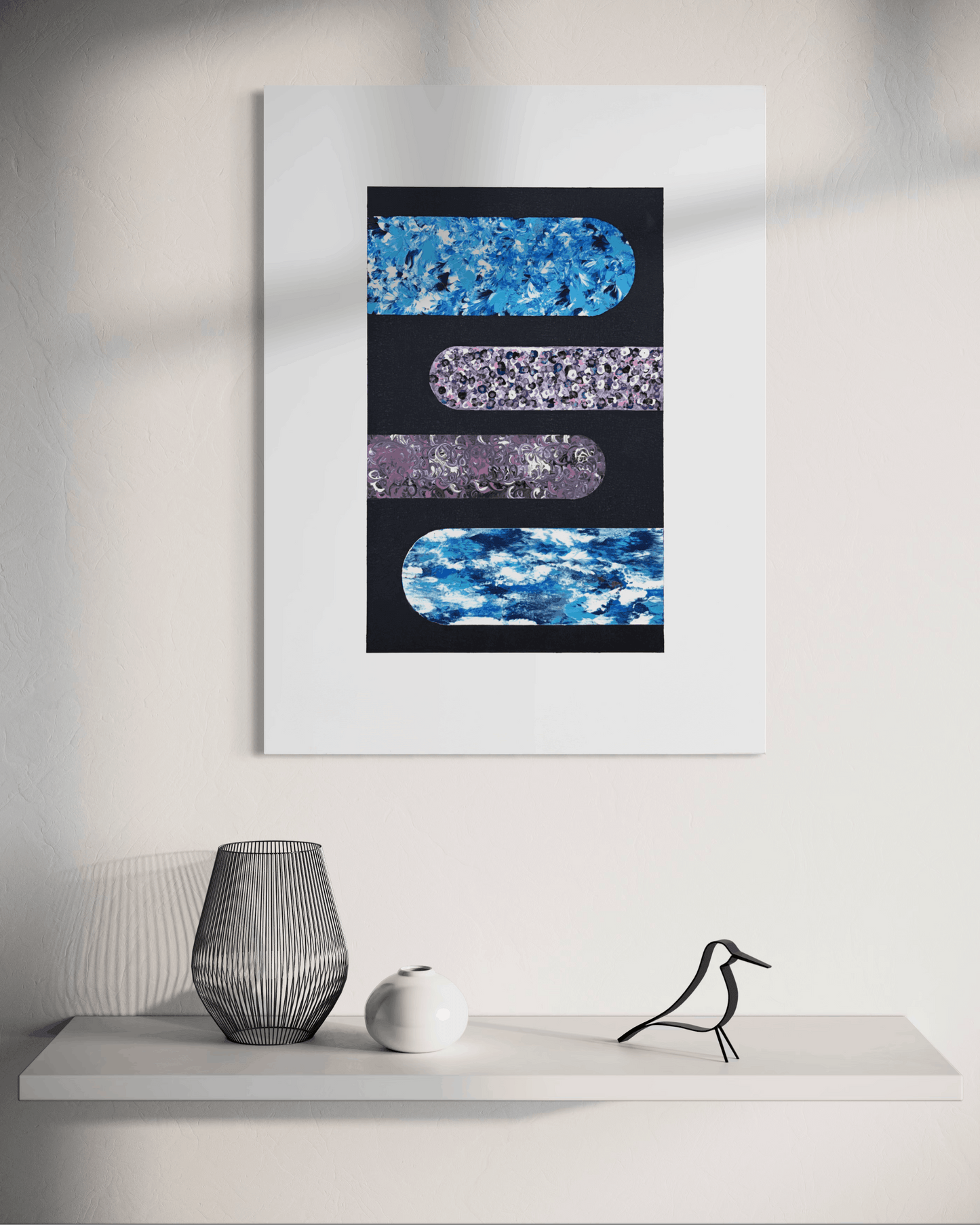 A framed abstract wall art print hangs on a white wall above a shelf. The artwork features three horizontal rounded rectangular bands in blue, purple, and blue patterns on a black background, surrounded by a wide white mat.