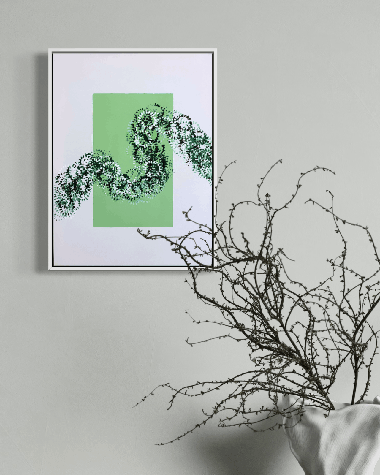 A framed abstract art print featuring a green square and a black-and-white snake-like pattern, displayed on a light gray wall next to a white vase with bare branches.