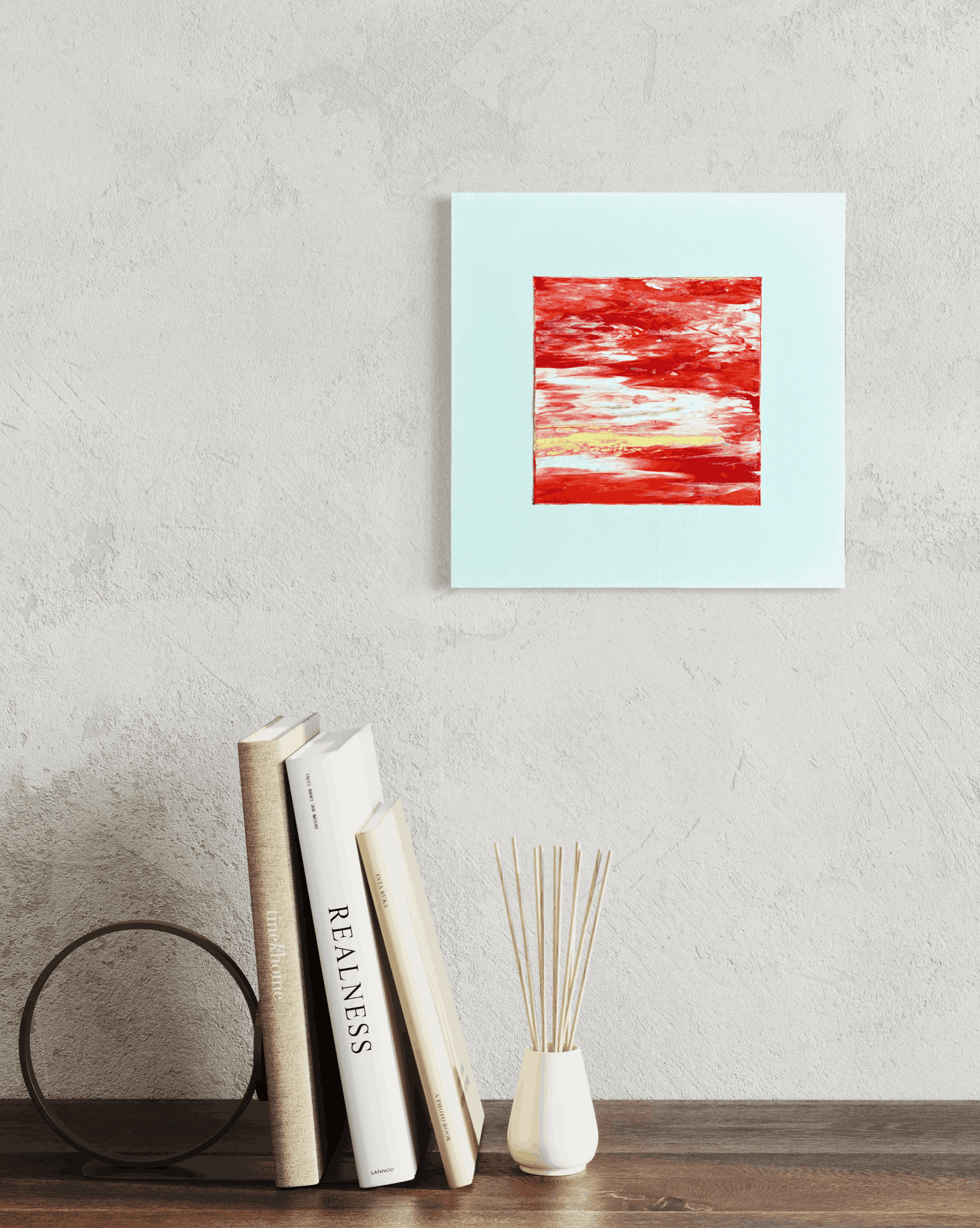 A square abstract wall art print with a light blue border and a textured red and white center, displayed on a light gray textured wall above a wooden shelf with books and a reed diffuser.