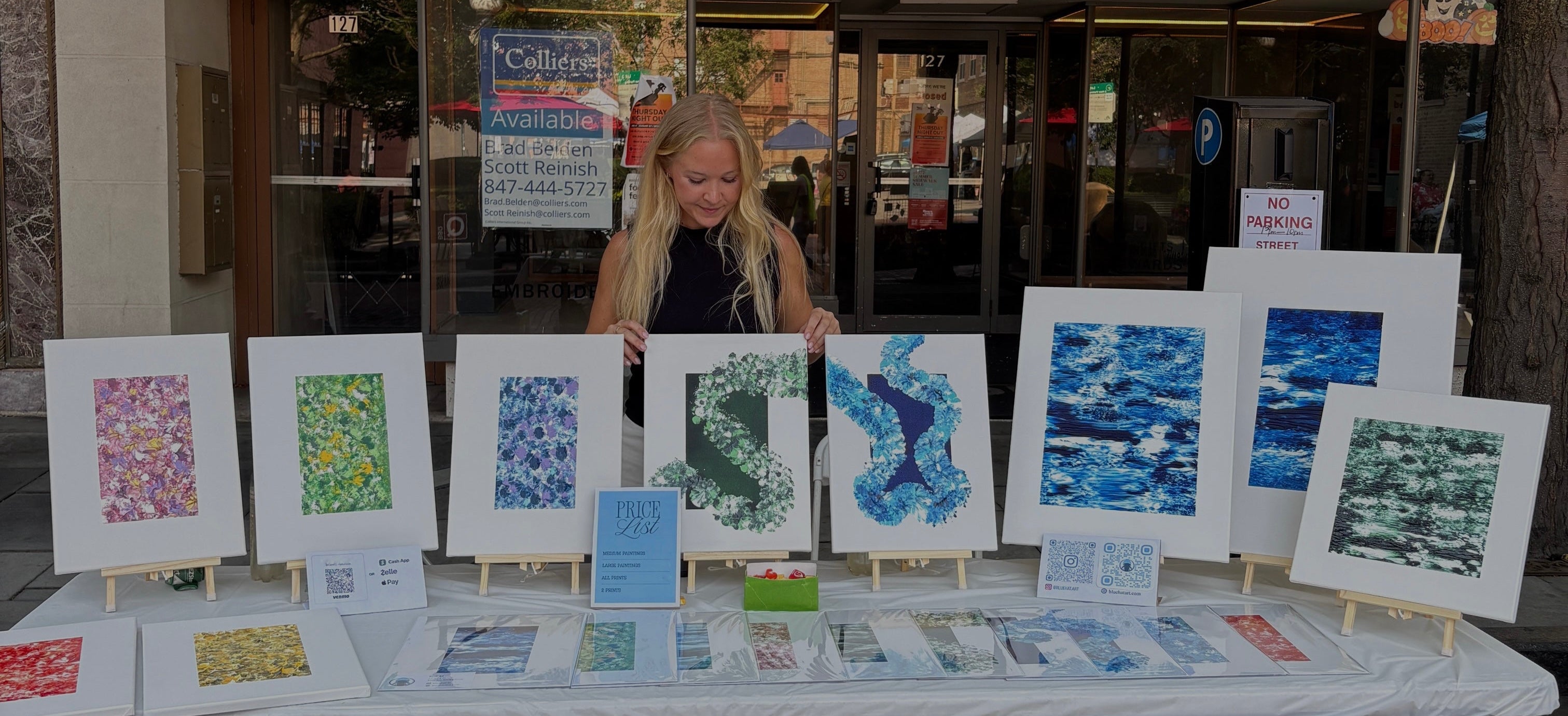 A woman stands behind a table displaying multiple colorful art prints and cards in front of a storefront with a sign reading 'Personalization House'.
