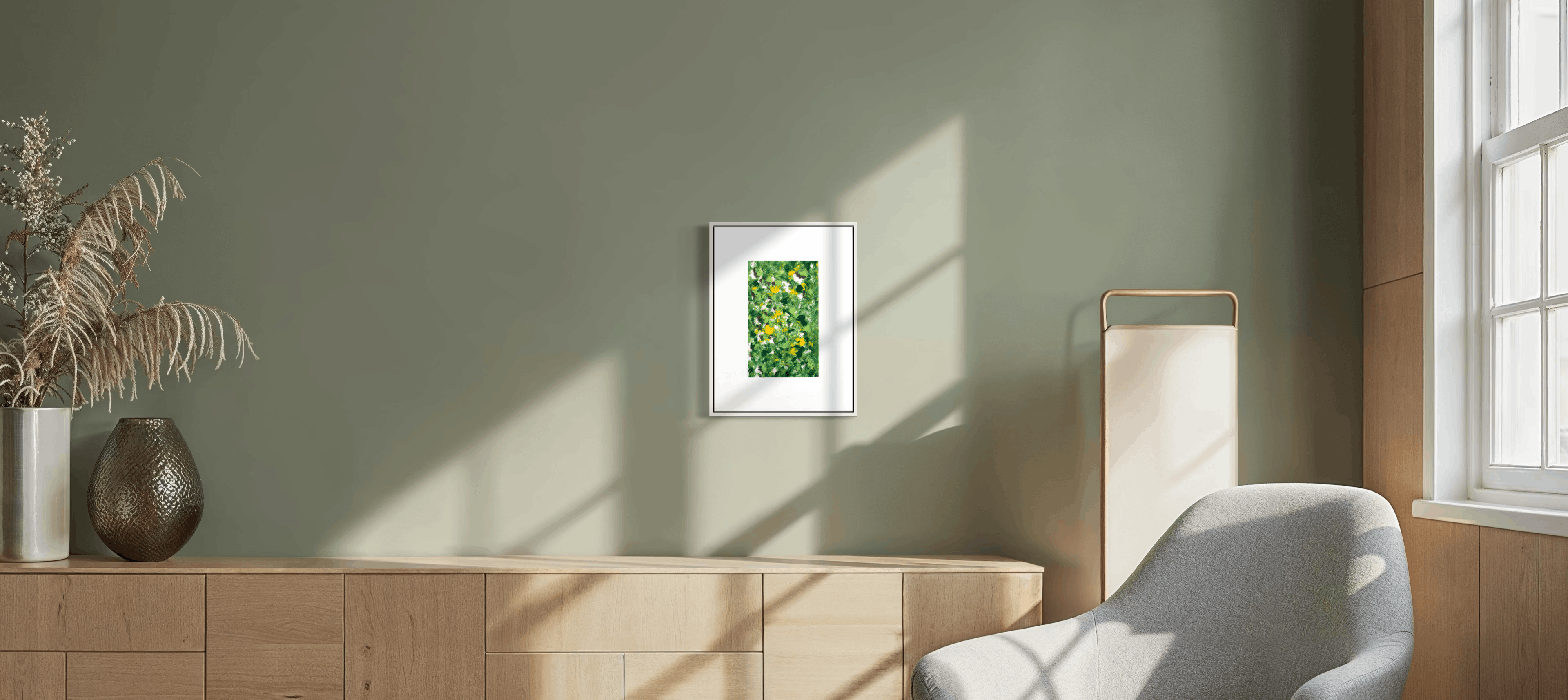 A small framed art print featuring a green leafy pattern, displayed on a sage green wall in a modern living room setting with a light wood console, a gray armchair, and warm sunlight casting shadows.