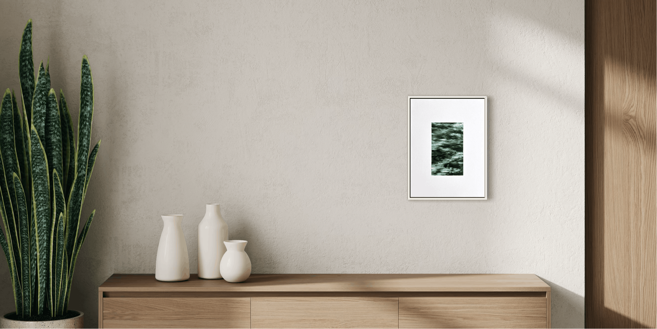 A framed abstract art print with green and black textured shapes on a white background, hanging on a textured beige wall above a light wood console table with white ceramic vases and a potted snake plant.