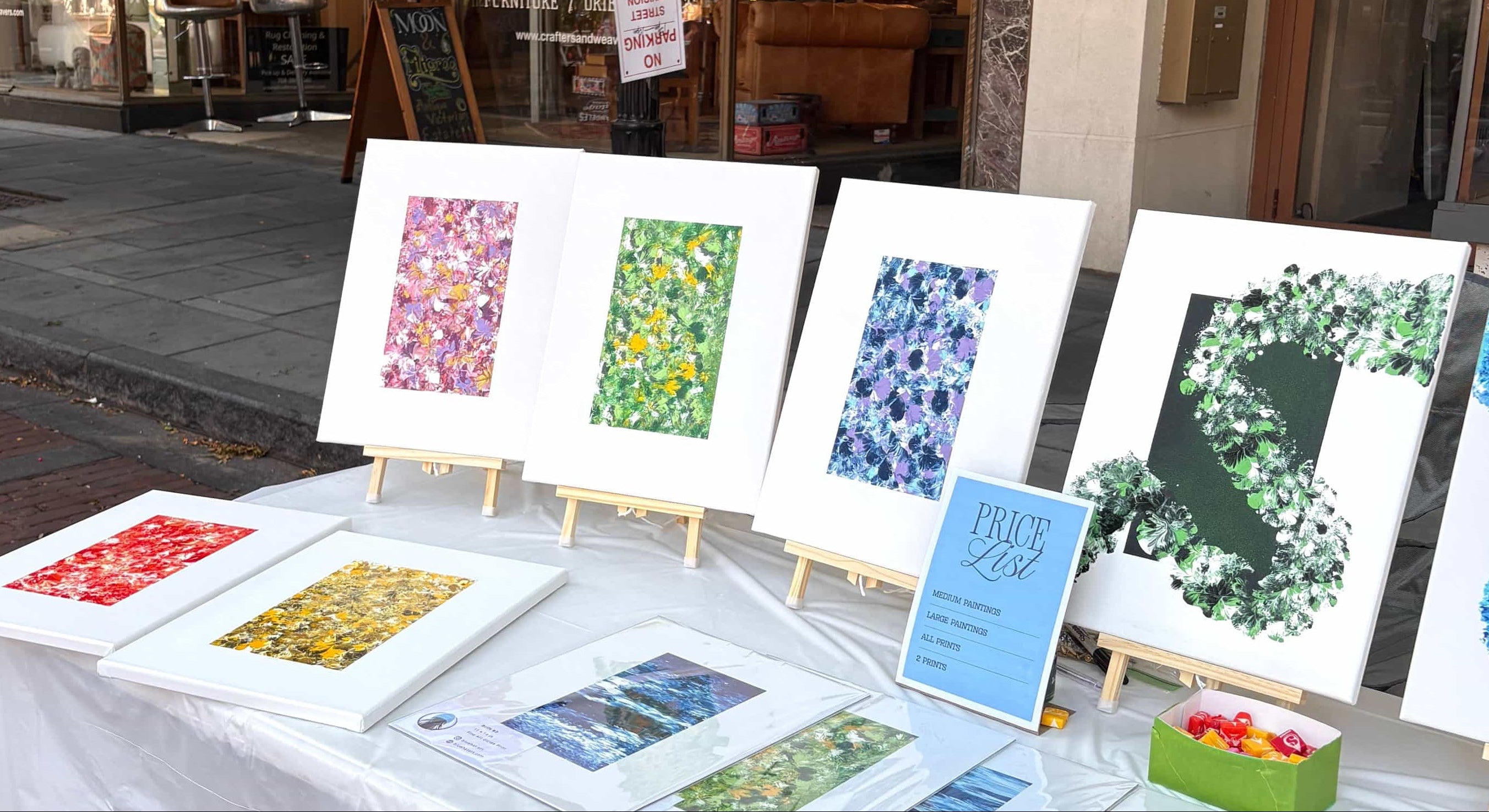 A street market display of framed art prints and paintings on easels and a table draped with a white cloth, set outdoors in front of a storefront.