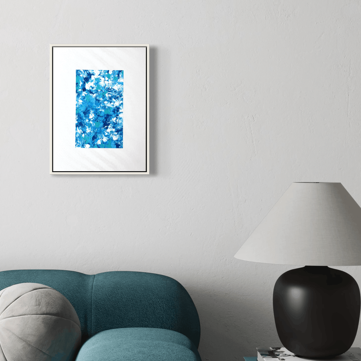 A framed abstract art print with vibrant blue and white brushstrokes hanging on a light gray wall above a teal sofa and next to a black table lamp.
