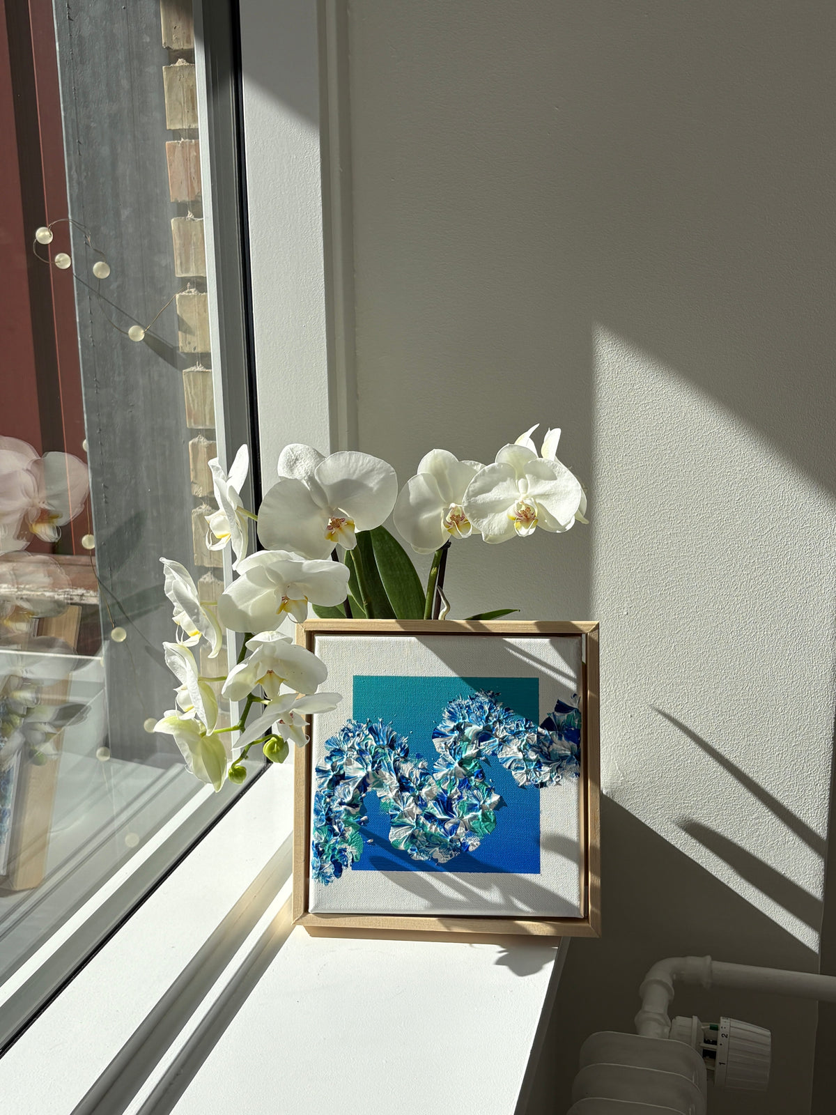 A framed floral art print featuring blue and white flowers on a textured blue background, styled on a white windowsill next to white orchids in a sunlit room.