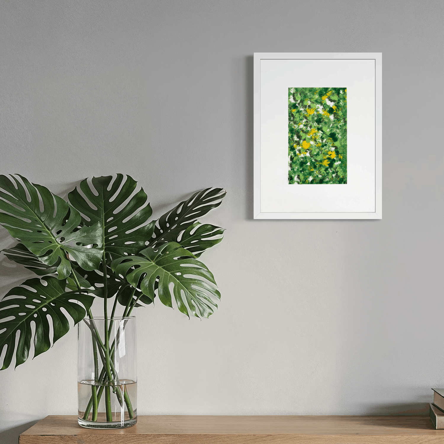 A framed art print featuring a green and yellow wildflower pattern, displayed on a gray wall above a wooden shelf with a large green monstera plant in a glass vase.
